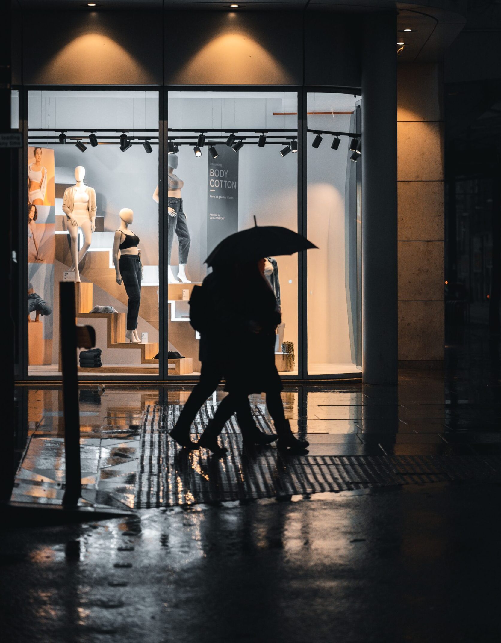 Silhouettes walking with umbrellas by a lit storefront on a rainy Manchester evening.